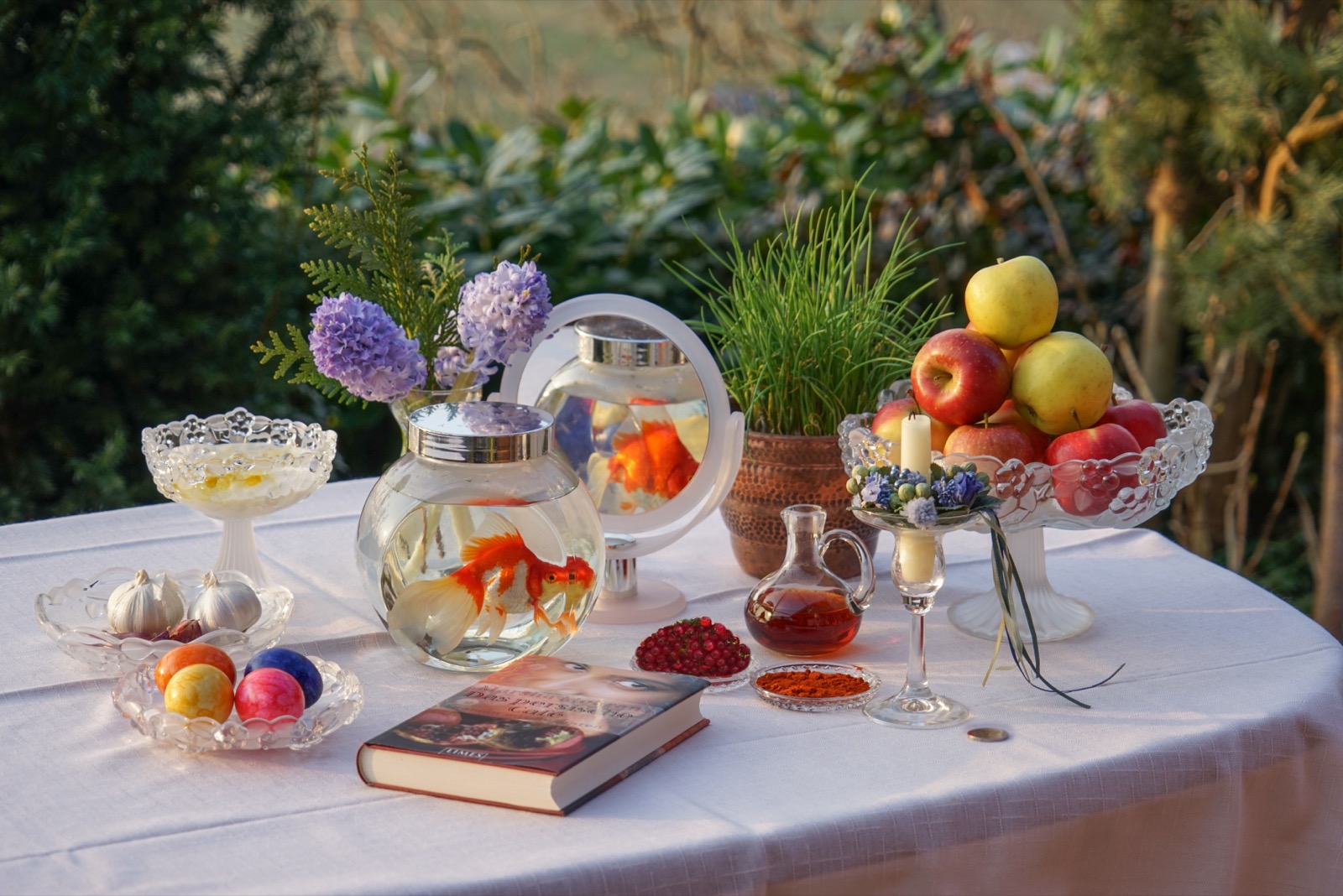 A full Nowruz haft-sin table outdoors with a goldfish bowl, mirror, hyacinths, apples, painted eggs and sabzeh