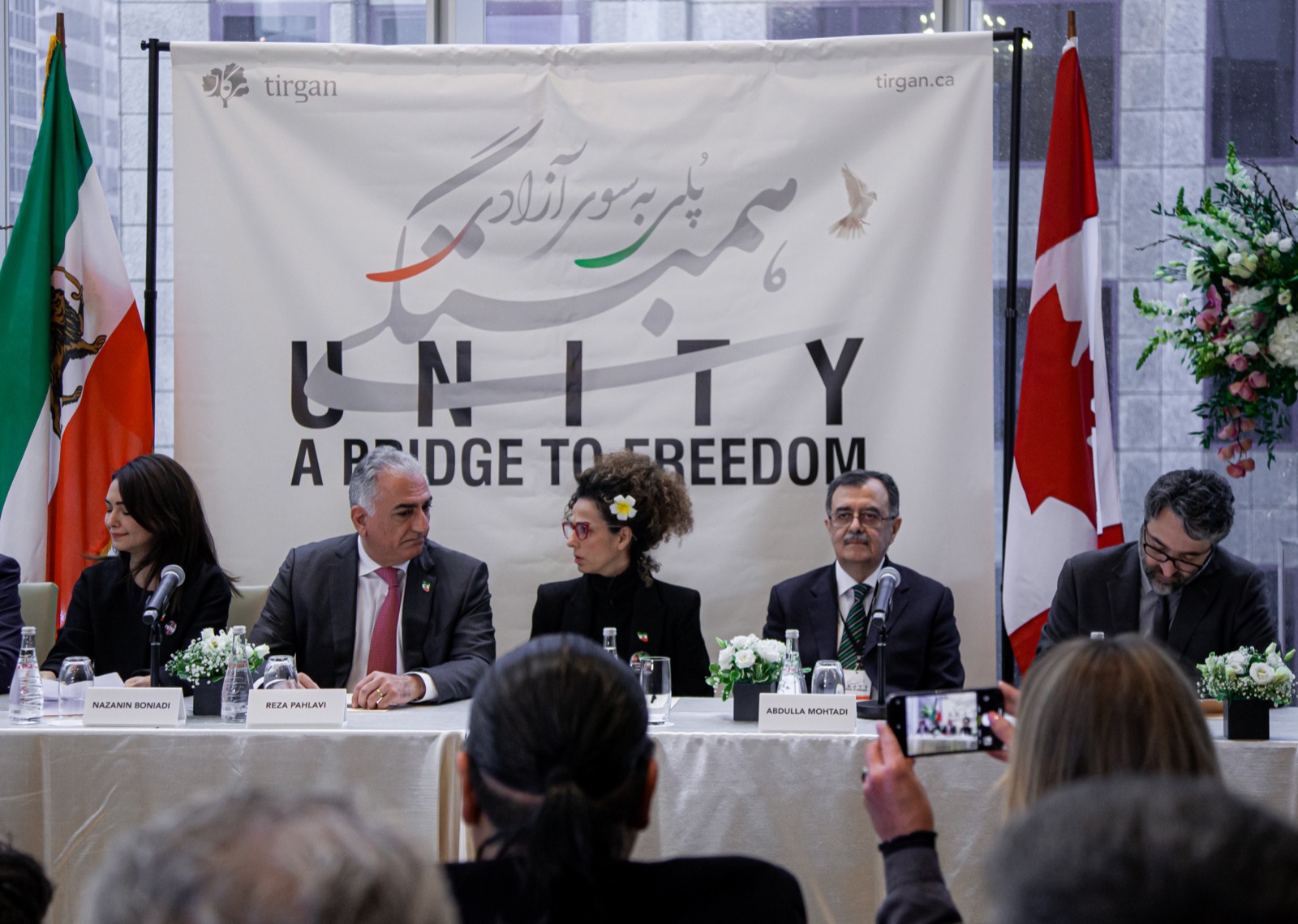 Five Iranian opposition figures seated behind name cards at a panel table with the Lion-and-Sun flag and Canadian flag, beneath a 'Unity — A Bridge to Freedom' banner, Toronto 2023